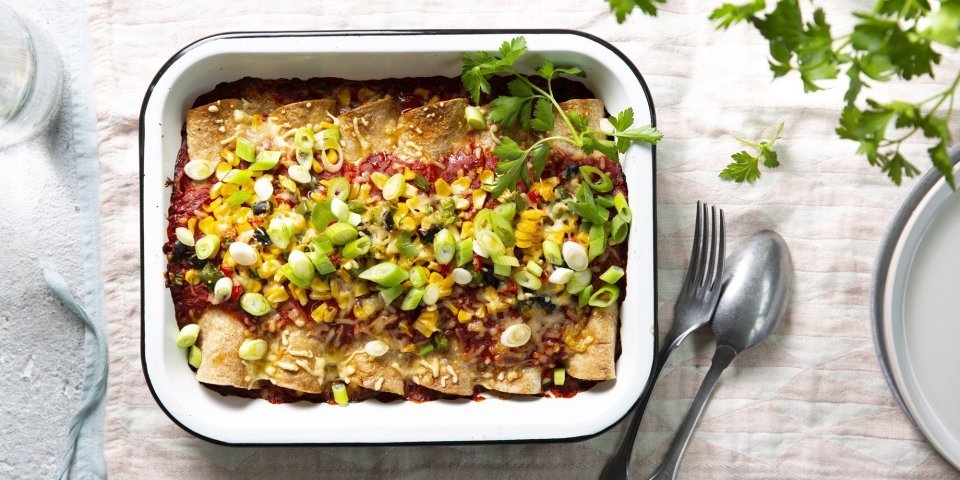 Vegetarische enchiladas Enchiladas liggen mooi op een rijtje in een witte ovenschotel. Hierop werd tomatensaus geserveerd en gemalen kaas, in de oven. Versgesneden lente-uitjes werden als afwerking toegevoegd aan het gerecht.