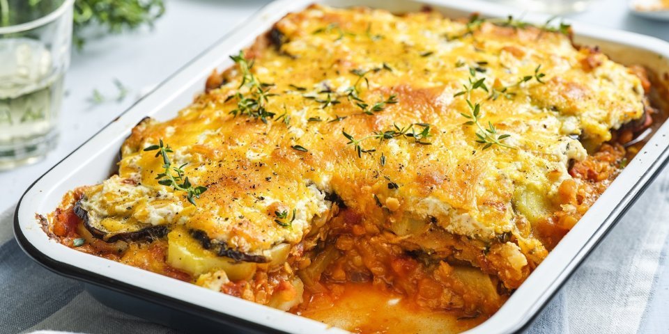 Er staat een grote schotel met vegetarische moussaka op tafel. Er is al een portie uitgeschept en op een bordje geserveerd. De moussaka is afgewerkt met verse tijmblaadjes. Op tafel staan ook nog 2 glazen en het plantje verse tijm.