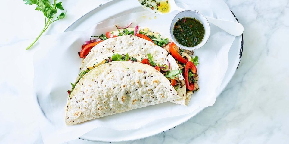 Vegetarische quesadilla gevuld met geroosterde knolselder, champignons, paprika en Brugge Oud. 2 stuks liggen gepresenteerd op een ovaal wit bord, samen met de vinaigrette in een potje apart. Takje verse koriander ligt er langs voor extra groen en sfeer aan het beeld mee te geven.