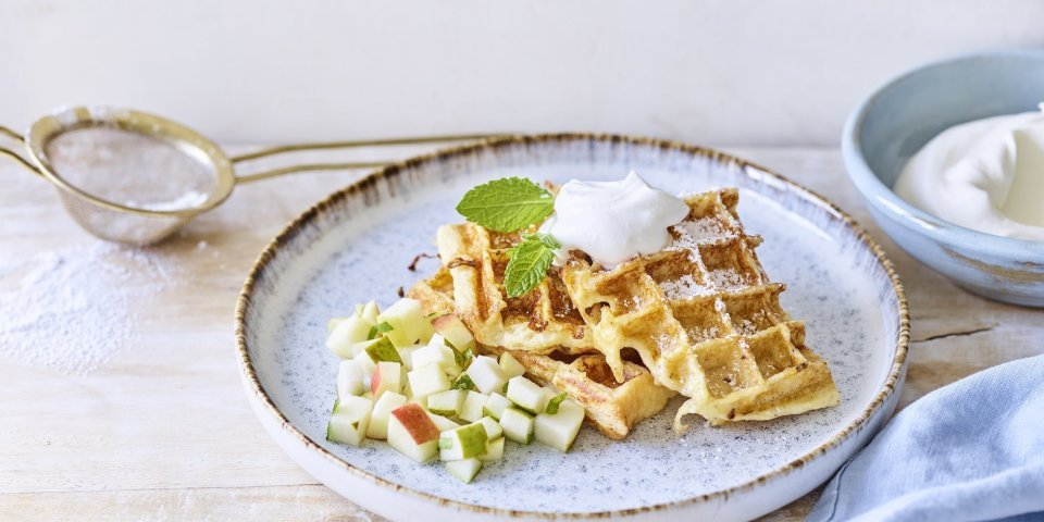 Verloren brood-wafeltjes met appel & peer Wafeltjes van verloren brood liggen op een licht grijs bordje. De stukjes fruit van appel en peer liggen onder de wafeltjes. We gaven ook een toefje slagroom op de wafels voor extra smaak. Er ligt een blauwe servette en klein zeefje met bloemsuiker langs het bord.