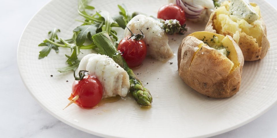 Een spies van visrolletjes, groene asperges en kerstomaten ligt op een wit bord. Ze worden geserveerd met een goudgele gepofte aardappel en natuurlijk een klompje boter erin. 