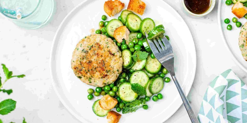 Visburger van heek met komkommer en erwtjessalade Een burger van vis? Yep, dit is er eentje van heek met een groene salade van erwtjes en komkommer. Langs het bord staat een klein sauspotje met de vinaigrette om zelf toe te voegen.