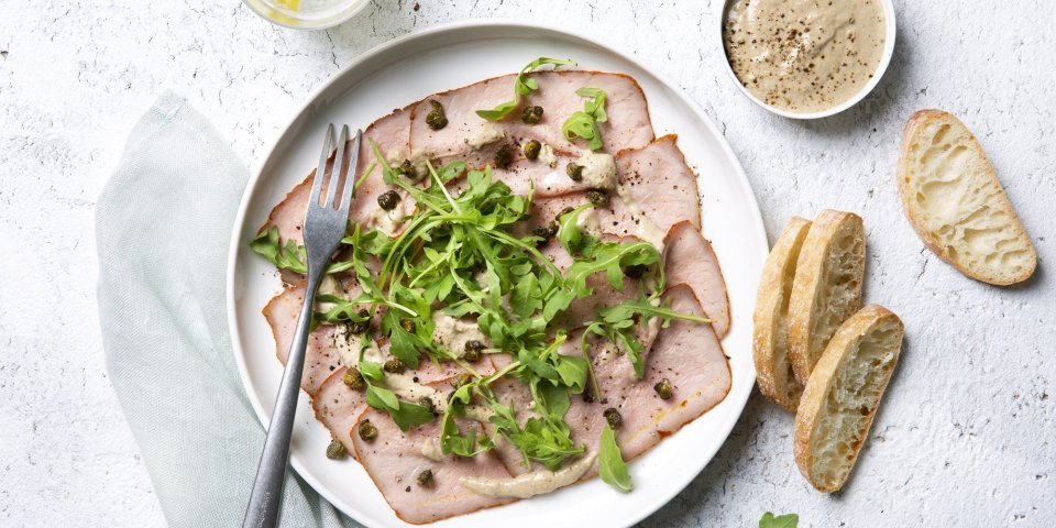 Deze vitello sgombro ligt op een wit bord, heerlijk afgewerkt met de koude saus, kappertjes en rucola. Er liggen ook enkele stukjes brood langs het bord.