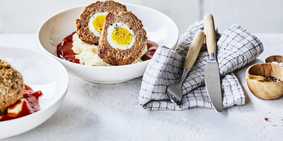 In een diep wit bord ligt de aardappelpuree onderaan met verse rode tomatensaus en daar bovenop de doormidden gesneden vogelnestjes. Er ligt een handdoek langs met blauwe ruitjes, hierop het bestek met houten handvat. 