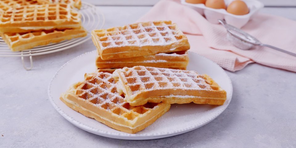 Een stappeltje klaargemaakte wafels ligt op een groot wit bord. Bloemsuiker is rijkelijk op de wafels gestrooid. In de achtergrond zie je nog enkele eitjes liggen en een roze linnen servette.