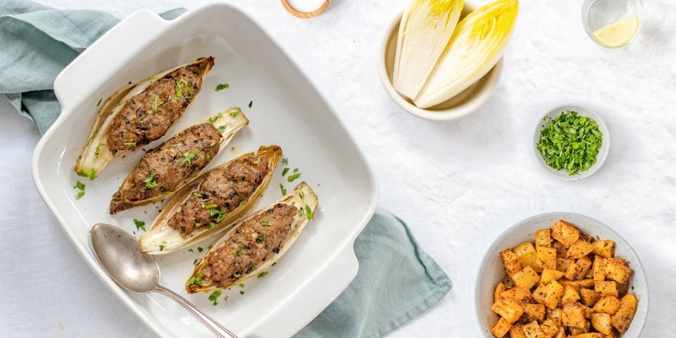 Gevulde kroppen witloof met kruidig gehakt liggen in een ovenschotel. De schotel staat op een lichtblauwe doek met kruidige aardappelen in kommetje langs. 