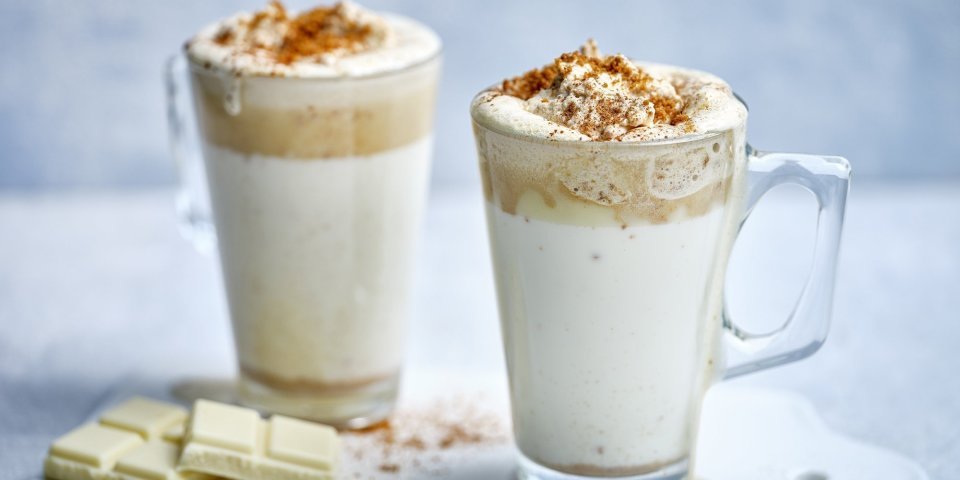 Twee hoge glazen tassen staan op het aanrecht klaar om te serveren. Ze zijn gevuld met witte chocolademelk, een goeie toef speculaasroom er bovenop en wat speculaaskruim. Er ligt een stukje witte chocolade naast. 