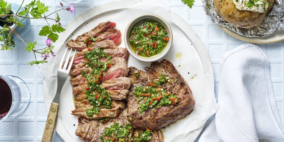 Het heerlijk stukje vlees ligt op een groot wit rond bord. Het al voor de helf in reepjes gesneden en bedruppeld met de Aziatische chimichurri. Er ligt een gepofte aardappel langs en twee glaasjes rode wijn. 