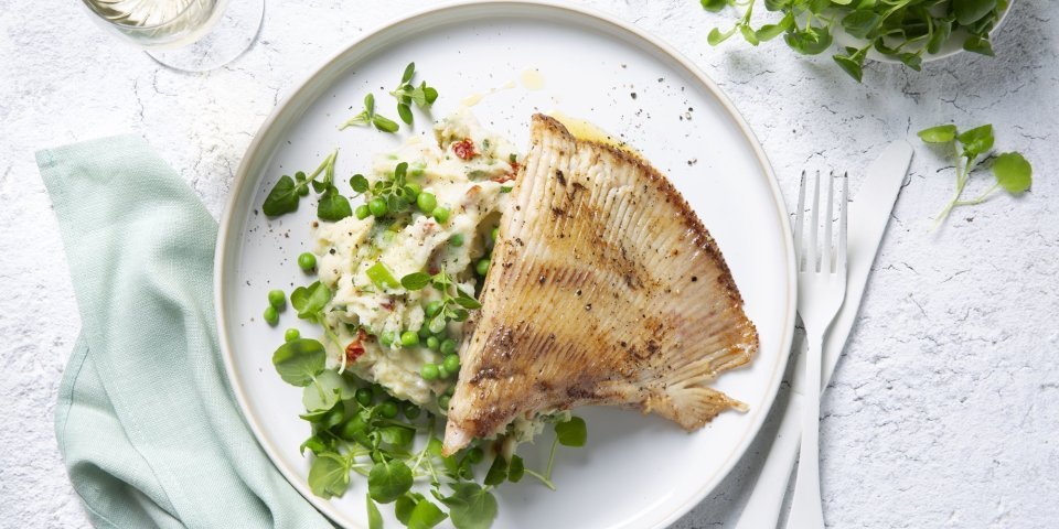 Zuiderse puree met rog Een wit bord gevuld met een frisse schep van de puree boordevol extra groentjes, denk maar aan verse lente-uitjes, zongedroogde tomaatjes, waterkers, kruiden enz. Hierop werd de gebakken rogvleugel gelegd die mooi krokant is gebakken. Naast het bord ligt ook nog een lichtblauwe servette, kommetje met extra waterkers en een glaasje wijn. Smakelijk!