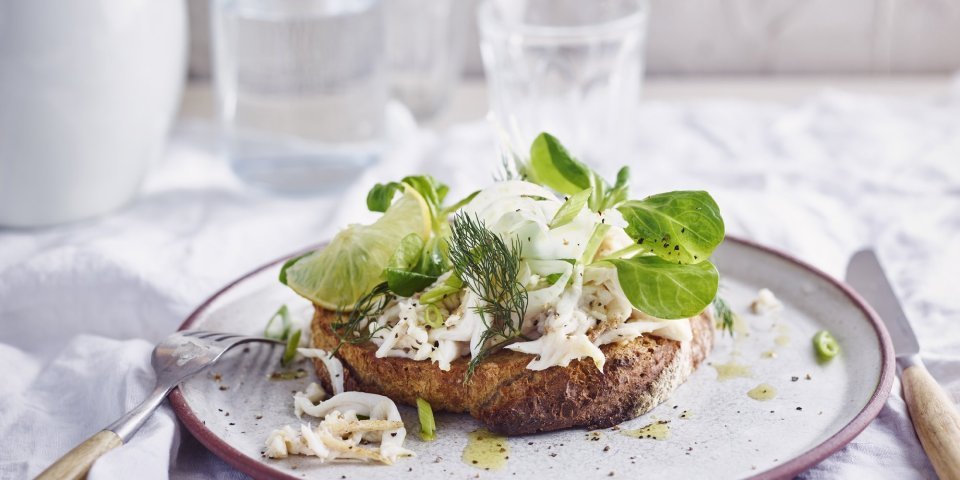 Zuurdesemtoast met vissla van rog, citroen en venkel Een heerlijke belegde toast van zuurdesem ligt op een wit bordje met rood-paarse rand. De toast is belegd met vissla van rog, citroen en venkel.