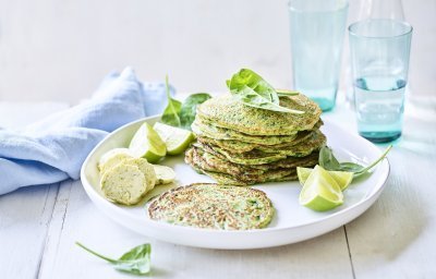 De pannenkoekjes van aardappel en spinazie liggen op een wit bord gestapeld. Er liggen blaadjes verse spinazie langs en partjes limoen. Op de witte houten tafel staan ook twee licht gekleurde blauwe glazen met water gevuld, een waterkan en een blauw linnen servette. 