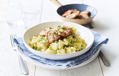 Een rijkgevulde aardappelsalade is opgediend in een diep wit bord. Hier bovenop ligt het krokant gebakken spek. De overige stukjes spek liggen nog in het bakpannetje en staat bovenaan het bord in beeld. 