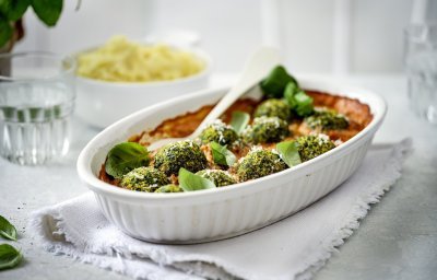 Een witte ovenschotel staat op tafel op een dikkere keukenhanddoek. De ovenschotel is gevuld met rode paprikasaus en hierop liggen de groene vegetarische balletjes van boerenkool. In de verte zie je ook nog een kommetje puree, klaar om aan tafel te gaan dus!