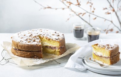 Een biscuit met vanillepudding en slagroom staat op tafel op een taartrooster. Er is al een stukje aangesneden om de lijnen duidelijk te laten zien van de pudding en slagroom. Koffie uitgeschonken, vorkje prikken en aanvallen die taart. 