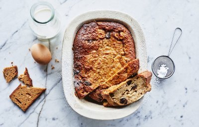 Een heerlijke homemade broodpudding staat op een marmer aanrecht in een witte ovenschaal. Hij is al aangesneden en een stukje uit de vorm genomen. Om de foto meer boddy te geven ligt er een eitje, flesje melk, stukje peperkoek en bloemsuiker naast.