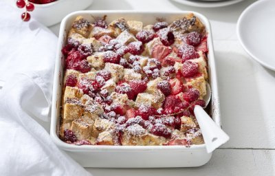 Op een witte houten tafel staat een rechthoekige ovenschotel met hierin de broodpudding klaargemaakt. Hij oogt zeer mooi door de toevoeging van het rode fruit en de afwerking met bloemsuiker. Er staan bordjes langs de ovenschotel om onmiddelijk op te scheppen en eventueel nog wat extra rood fruit te nemen. 