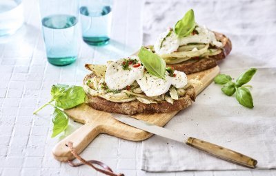 Een rijkbelegde toast met venkel en mozzarella liggen op een houten plank. Boven deze plank staan twee blauwe glazen gevuld met water. Het gerechtje is afgewerkt met enkele basilicumblaadjes. 