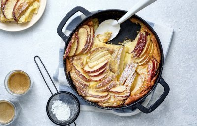 Een clafoutis van appelstukkjes staat op het aanrecht in een zwarte fonte schaal. Er is al een portie uitgeschept en staat op een bordje naar de pot. Er is ook bloemsuiker over het dessert gestrooid en de koffie is al uitgeschonken. 