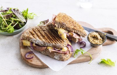 De croque ligt op een houten plankje aangesneden. De kaas en rode ui zien er heerlijk uit. In de achtergrond zie je ook nog wat gemengde sla en een potje graantjesmosterd. 