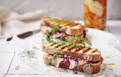 Twee croques liggen op het aanrecht in de keuken op een wit boterpapiertje. In de achtergrond staan de gepekelde groentjes (wortel en komkommer) in een bokaal. De tosti's zijn mooi gegrild in streepjes.