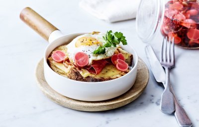 Croute au fromage met Averbode kaas in een witte pan, spiegeleitje er bovenop gepresenteerd met een takje verse peterselie. Op de achtergrond staan gepekelde radijzen in glazen pot.