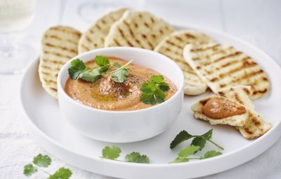 Homemade paprikadip zit in een wit schaaltjes. Het naanbrood ligt er langs op een wit bord. Het gercht is afgewerkt met korianderblaadjes.