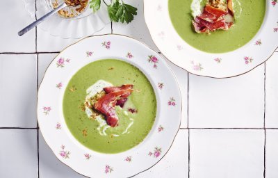 2 porties erwtensoep staan klaar op het werkblad van de keuken. De erwtensoep heeft een mooie groene kleur die mooi contrast geeft in de witte borden. De soep is afgewerkt met peterselieyoghurt, krokante ham en gefruite uitjes.