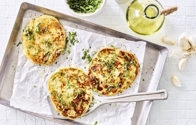 Drie broodjes zijn gebakken, en wat voor een! Flatbread gemaakt van een restje aardappelpuree liggen te blinken op een grijze bakplaat met wit boterpapiertje op. Verse kruiden liggen klaar om af te werken. 