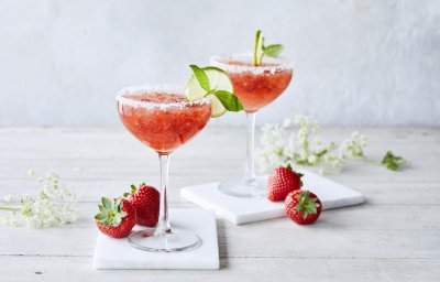 Cocktail time! Wij serveren een frozen daiquiri met verse aardbeien en basilicum. We presenteren hem in een wijnglaasje met suikerrandje, schijfje limoen en basilicumblaadje. Er liggen enkele aardbeien langs samen met witte bloemetjes. Schol! 