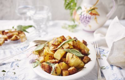 Gebakken aardappelen liggen in een groot wit bord op een gedekte tafel. De verse salieblaadjes geven de krokante aardappelstukjes extra kleur. 
