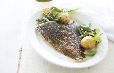 De gebakken pladijs op de graat ligt op een groot wit ovaal bord. Wij serveren hem met boontje langs en enkele hasselback potatoes. Het bord staat op een witte houten tafel en de salsa verde staat apart in een potje boven het bord.  