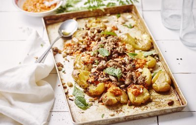 Op een ovenschaal liggen krokante crushed potatoes met homemade romesco saus bovenop en afgewerkt met lamsgehakt. De overige romesco saus staat bovenaan voor wie wat extra wil. Er ligt een witte servette op het aanrecht van witte Marokaanse tegeltjes. 