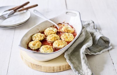 Witte ovenschotel staat op een houten snijplankje op tafel. De ovenschotel is gevuld met de rode tomatensaus waarin de champignons mooi in geschikt zijn. Achteraan de foto zie je bordjes staan met bestek om aan de slag te gaan. 