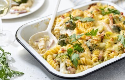 Op tafel staat een ovenschotel met hierin de pasta, prei, broccoli en kaas. Hij is gegratineerd in de oven, wat een mooi goud korstje geeft en afgewerkt met verse peterselie. Er is al een portie uitgeschept en het bordje staat boven de shotel. 