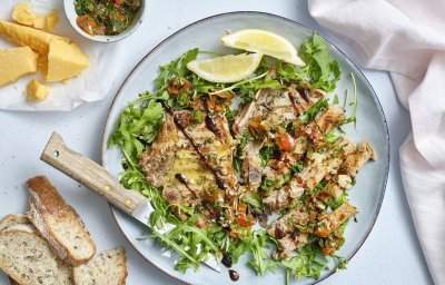 Op een groot blauw/grijs bord ligt eerst een salade van rucola waarop de mooi gegrilde kotelet van kalfsvlees op ligt. De kotelet is in reepjes gesneden waarop de gremolata van tomaten en de oude kaas over gestrooid is. Naast het bord liggen de brokkelkaas en de rest van de gremolata. 