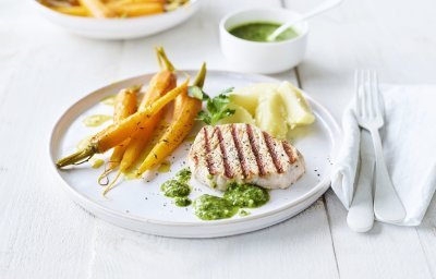 Een mooi in streepjes gegegrild stukje varkensvlees ligt op een wit bord, geserveerd met gegaarde wortels en gekookte aardappelen. We serveren geen saus bij dit stukje vlees, maar homemade pesto van peterselie en Brugge kaas. 