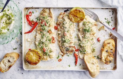 3 stukjes gegrilde vis liggen op een rechthoekige ovenschaal. De gegrilde citroen en chilipepers zorgen voor kleur in dit gerechtje, samen met de groene kruiden in de crumble en dip. 