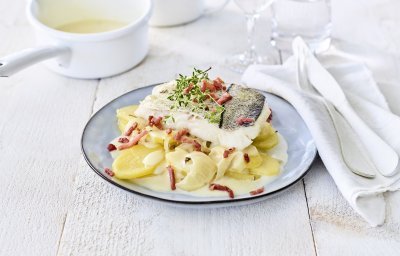 Schelvis ligt op een torentje aardappelen in een blauw bord. Het bordje staat klaar om aan te vallen op een houten witten tafel met wit bestek langs.