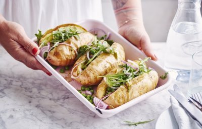 3 gepofte aardappelen met pesto, erwtjes en rucola liggen in een witte ovenschaal. De vrouw wil de ovenschaal plaatsen op tafel.