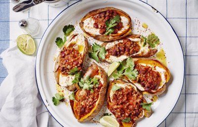 Een ronde ovenschaal met de gepofte zoete aardappelen staat op tafel. De gepofte aardappelen zijn gevuld met yoghurt en hierbovenop de chili sin carne, ze zijn afgewerkt met enkele blaadjes koriander en partjes limoen. De witte ovenschaal heeft een blauw randje en staat op een blauw geruite handdoek.