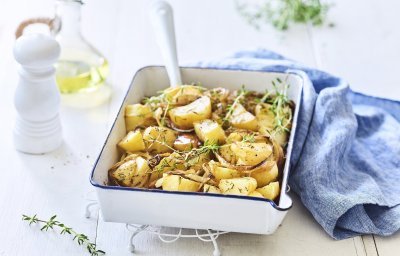 Een witte ovenschaal met donkerblauw randje staat op een keukenonderlegger op een witte houten tafel. Er staat een witte pepermolen en een glazen flesje met olijfolie langs. De verse tijm zorgt voor extra kleur bij de geroosterde aardappelen, er ligt ook nog een blauwe linnen servette langs de ovenschotel.