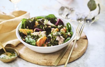 Een salade van bietjes, boerenkool en spinazie afgewerkt met noten en makreel. Deze feestsalade staat op een houten plank met de dressing in een apart gouden potje.