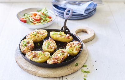 7 gevulde aardappelen liggen op een zwarte vuurvaste pan, ze zijn gevuld met aardappelkruim, kaas en spekblokjes. Er ligt een handdoek achteraan in beeld en een slaatje met tomaatjes. 