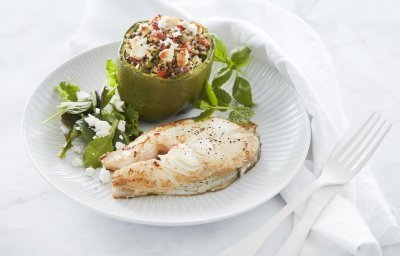 Een gebakken moot van schelvis ligt op een wit bord. Hij is geserveerd met een gevuld groene paprika. Deze is opgevuld met quinoa, tomaten- en komkommerstukjes en geitenkaasblokjes.