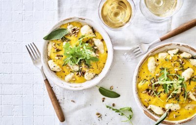 Gnocchi met pompoensaus en salie staan klaar op een tafel met wit tafelkleed. Bestek met houten afwerking ligt bij het bord, het gerechtje is afgewerkt met verse blaadjes salie en pitten. 