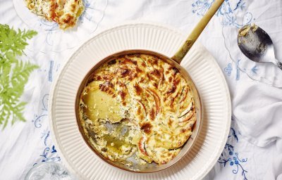 Een heerlijke gratin met groenten en kaas klaargemaakt in een ovenbestendige pan staat in het midden van de tafel. Er is al een portie uitgeschept op een bordje. Op tafel ligt een wit tafelkleed met enkele geborduurde blauwe bloemen. 