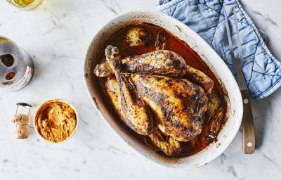 Een heerlijk gegrilde kip uit de oven ligt in een ovenshaal. Ze is heerlijk krokant gebakken en lekker sappig door de homemade kruidenboter. Ze staat op het marmere aanrecht met blauwe nap er langs. 