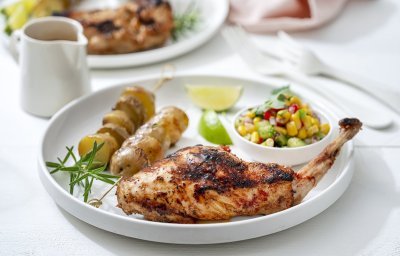 Een gegrilde konijnenbout ligt op een wit bord, geserveerd met een gemengde salade van maïs en paprika en een brochette van aardappel.