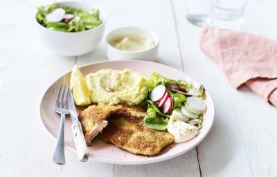 Een homemade shnitzel ligt op een pastelroze bord, samen met aardappelpuree, verse sla en zelfgemaakte mayonaise. Er is nog wat extra salade in een wit potje en mayonaise voor extra lekkers op te scheppen. 