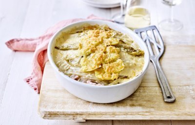 Er staat een ovenschotel op een houten plank, op de tafel. Er ligt een lepel naar de ovenschotel die gevuld is met een lasagne van witloof, ham, kaas en aardappelen. Er ligt ook een coraalroze servette bij.